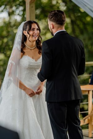 Smiling bride in lace gown holds grooms hands during outdoor wedding vows.
