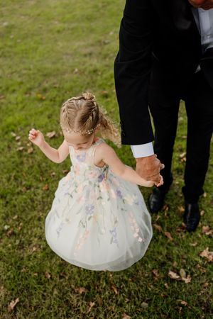 Adorable flower girl in a floral tulle dress holds hands with a man in a black suit.