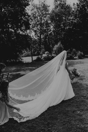 Black and white photo of a bride in a flowing gown with a long lace-trimmed veil billowing behind her outdoors.