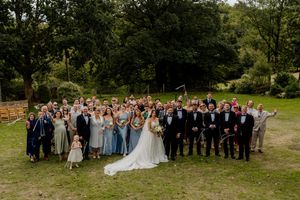 Large wedding party group photo on green lawn surrounded by lush trees.