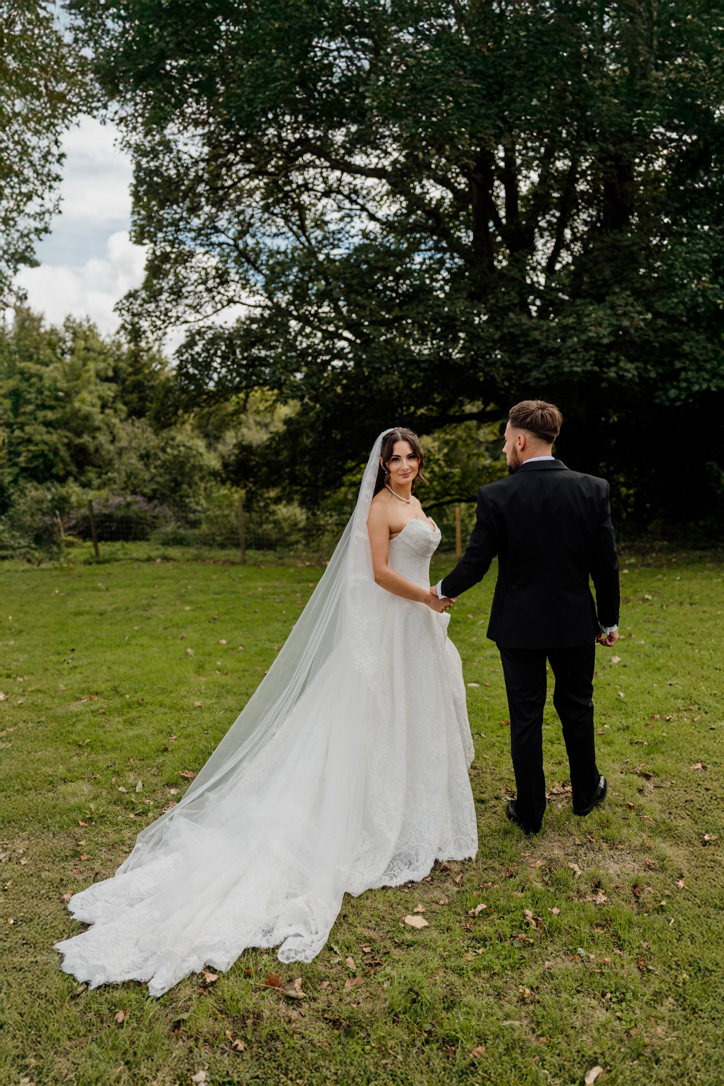 Bride in white gown with long veil holds grooms hand walking through lush green garden on wedding day.