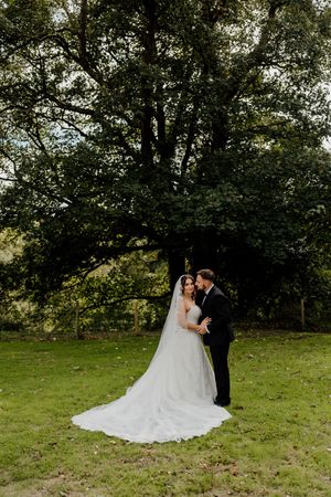 Bride and groom share romantic moment beneath large tree in garden setting.