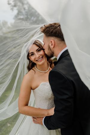 Groom kisses smiling bride wrapped in flowing veil outdoors.