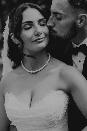 Black and white photo of groom kissing bride wearing pearl necklace and strapless gown.