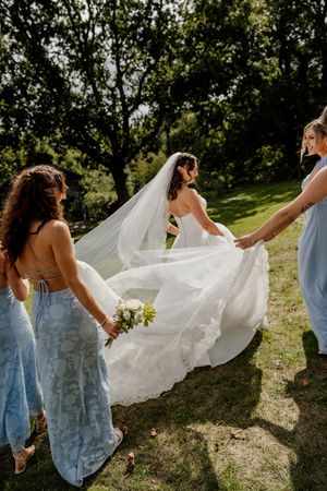 Bridesmaids in blue dresses help bride with train and veil outdoors.