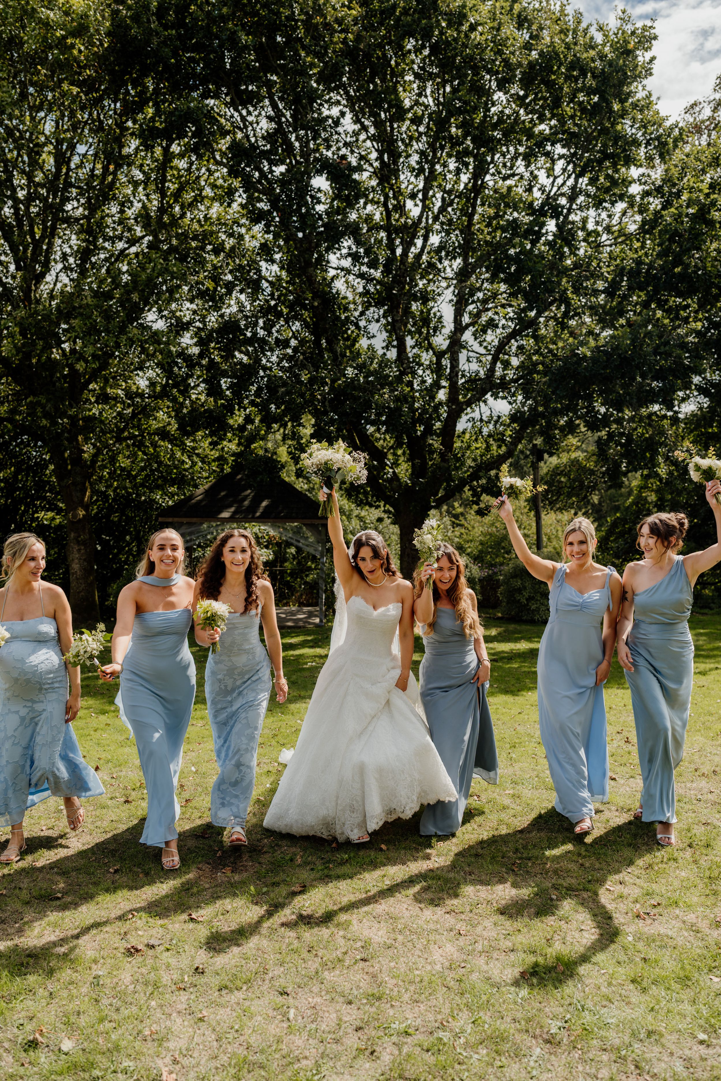 Bride and bridesmaids in blue dresses celebrate joyfully on sunny garden lawn.
