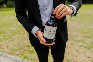 Groom in black tuxedo holding a bottle of Black Tot rum during an outdoor wedding celebration.