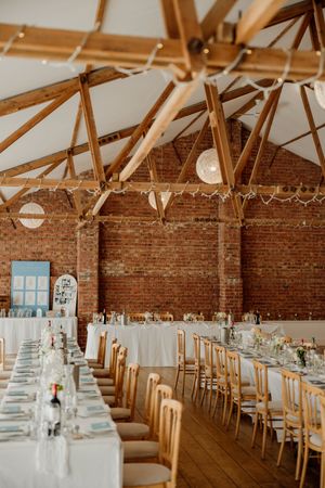 Rustic barn wedding venue with exposed brick walls, wooden beams, fairy lights, and long white-clothed reception tables with gold chairs.