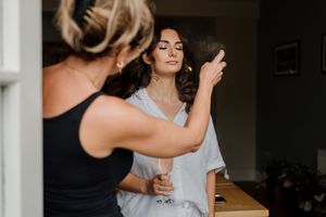 A hairstylist sprays a bride's elegant dark hair during wedding preparations in front of a mirror.