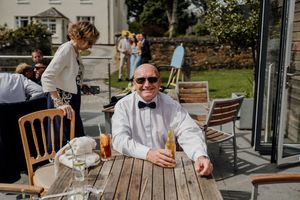 Smiling wedding guest in bow tie and sunglasses relaxing outdoors with drinks at wooden table.