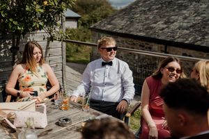 Wedding guests laughing and socializing outdoors at rustic stone venue on sunny day.