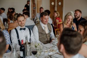 Two grooms smile at wedding reception table with guests, wine bottles, and floral centerpieces in bright venue.