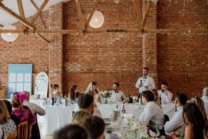 Wedding reception speech in rustic brick barn venue with string lights, white tables, and seated guests listening.