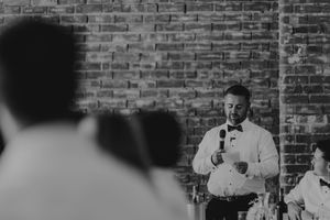 Man in white shirt giving a speech at a wedding reception, holding a microphone against a brick wall backdrop.
