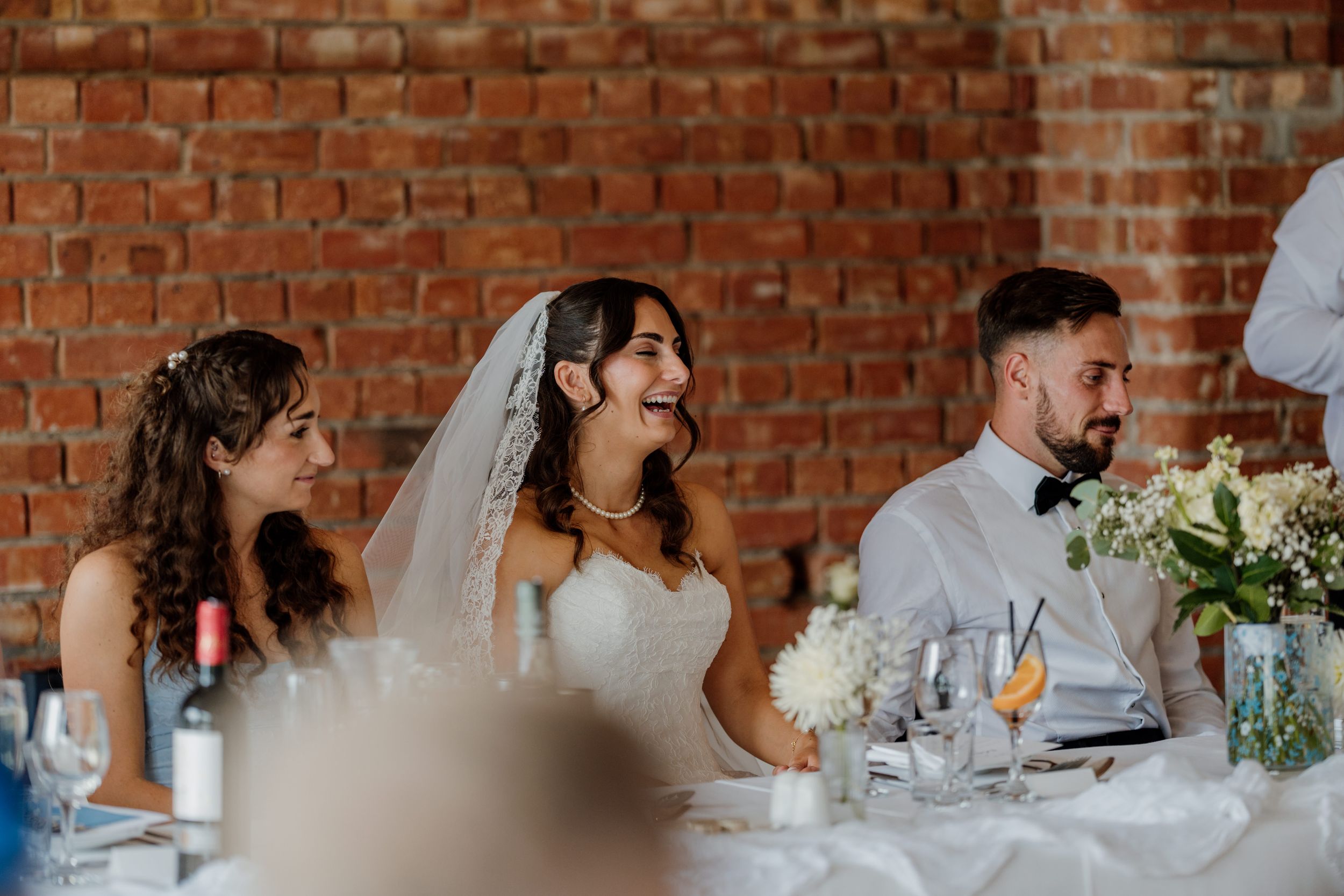 Bride laughing joyfully at wedding reception table with guests, brick wall background.