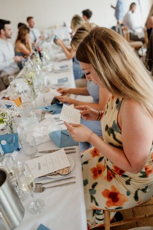 Wedding guest in floral dress reading a card at an elegantly set reception table with blue napkins.