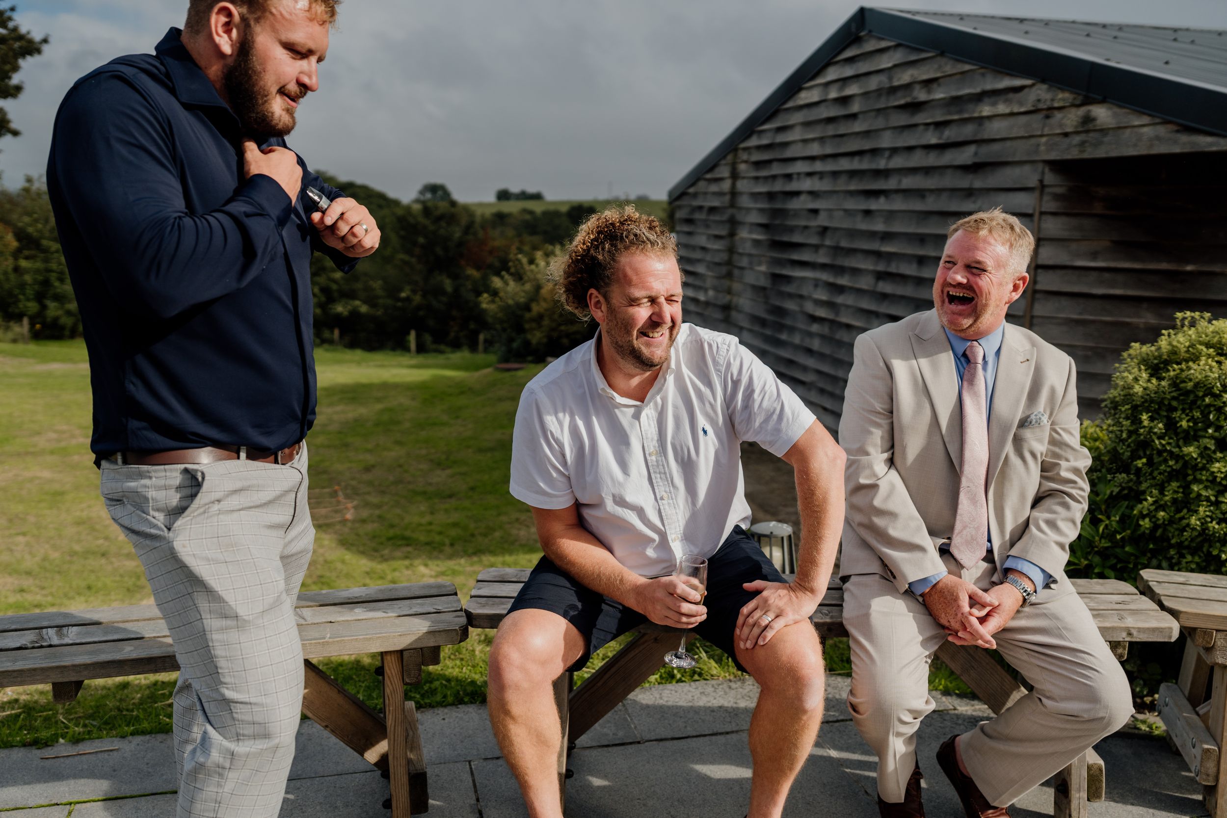 Three men laughing outdoors near wooden benches at a countryside wedding venue on a sunny day.