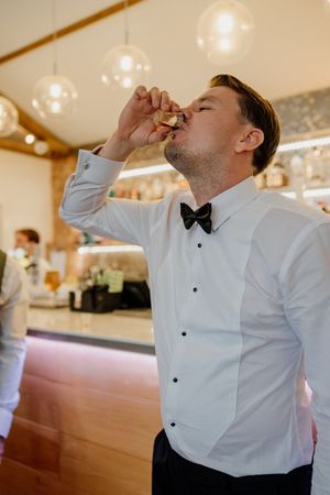 A groom in a white dress shirt and black bow tie takes a celebratory shot at a wedding bar.