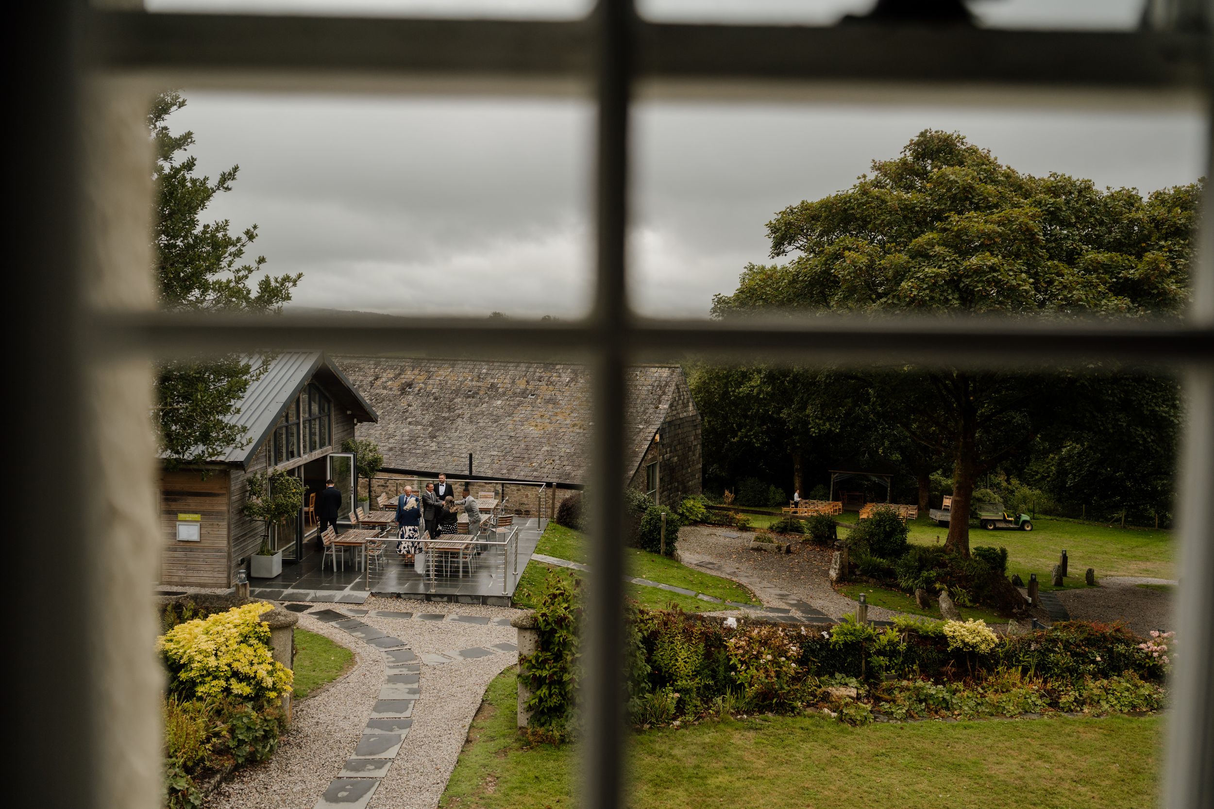 View through a window of a rustic barn venue with manicured gardens and stone pathways on an overcast day.
