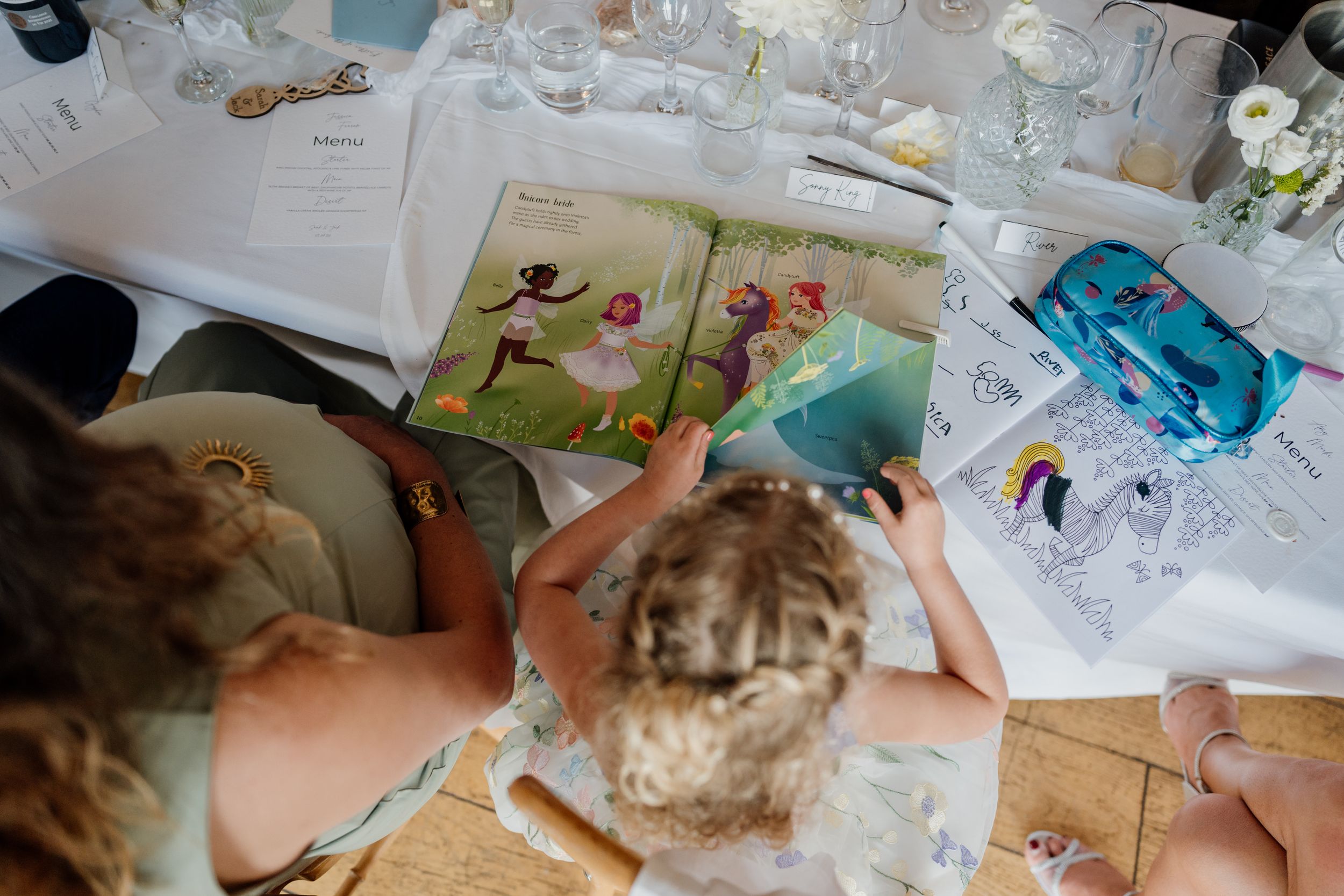 Children reading a colorful illustrated book at a decorated wedding reception table with coloring pages nearby.