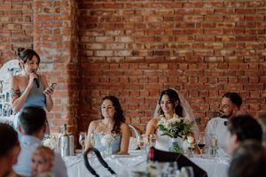 Wedding reception speech at a rustic brick wall venue with bride, bridesmaid, and guests seated at decorated tables.