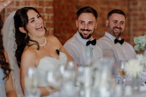 Bride and groom laughing joyfully at wedding reception table with groomsmen in white shirts and bow ties.