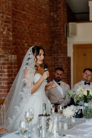 Bride in lace veil gives speech holding microphone at wedding reception with brick wall backdrop.