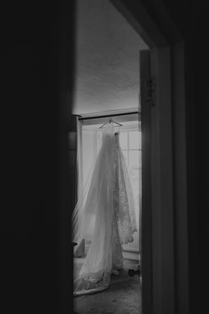 A black and white photo of an elegant lace wedding dress hanging in a window.