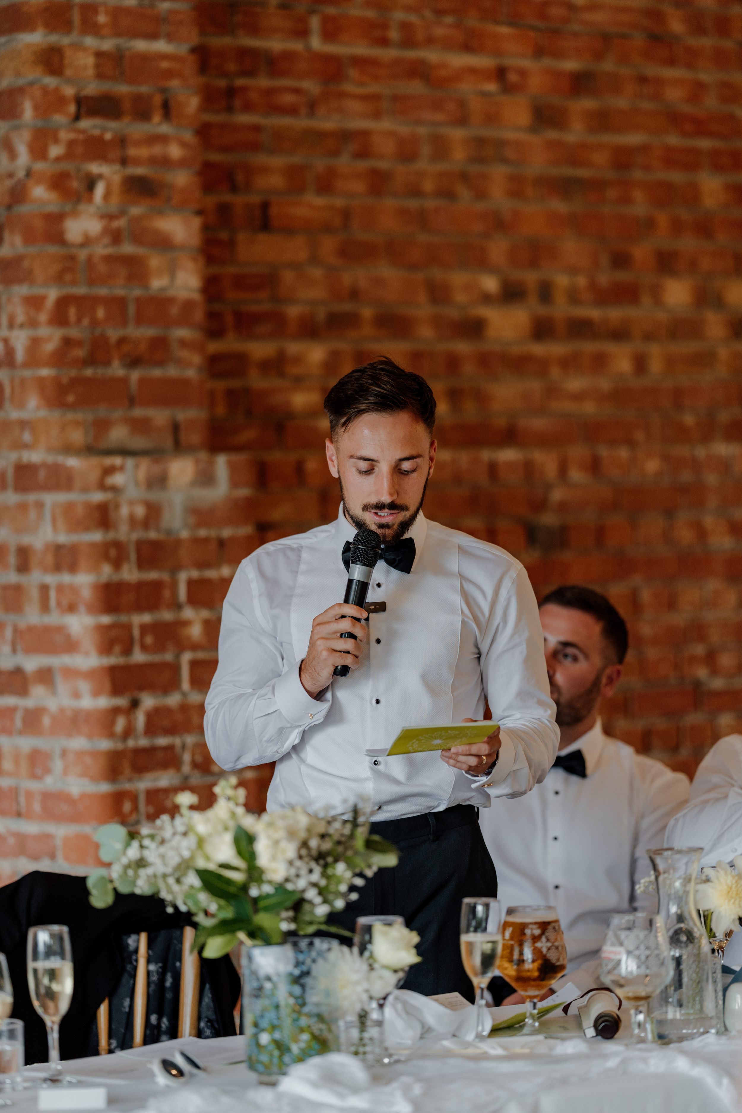 Groom delivering a wedding speech at a reception, holding a microphone and notes, with floral centerpieces and brick wall backdrop.