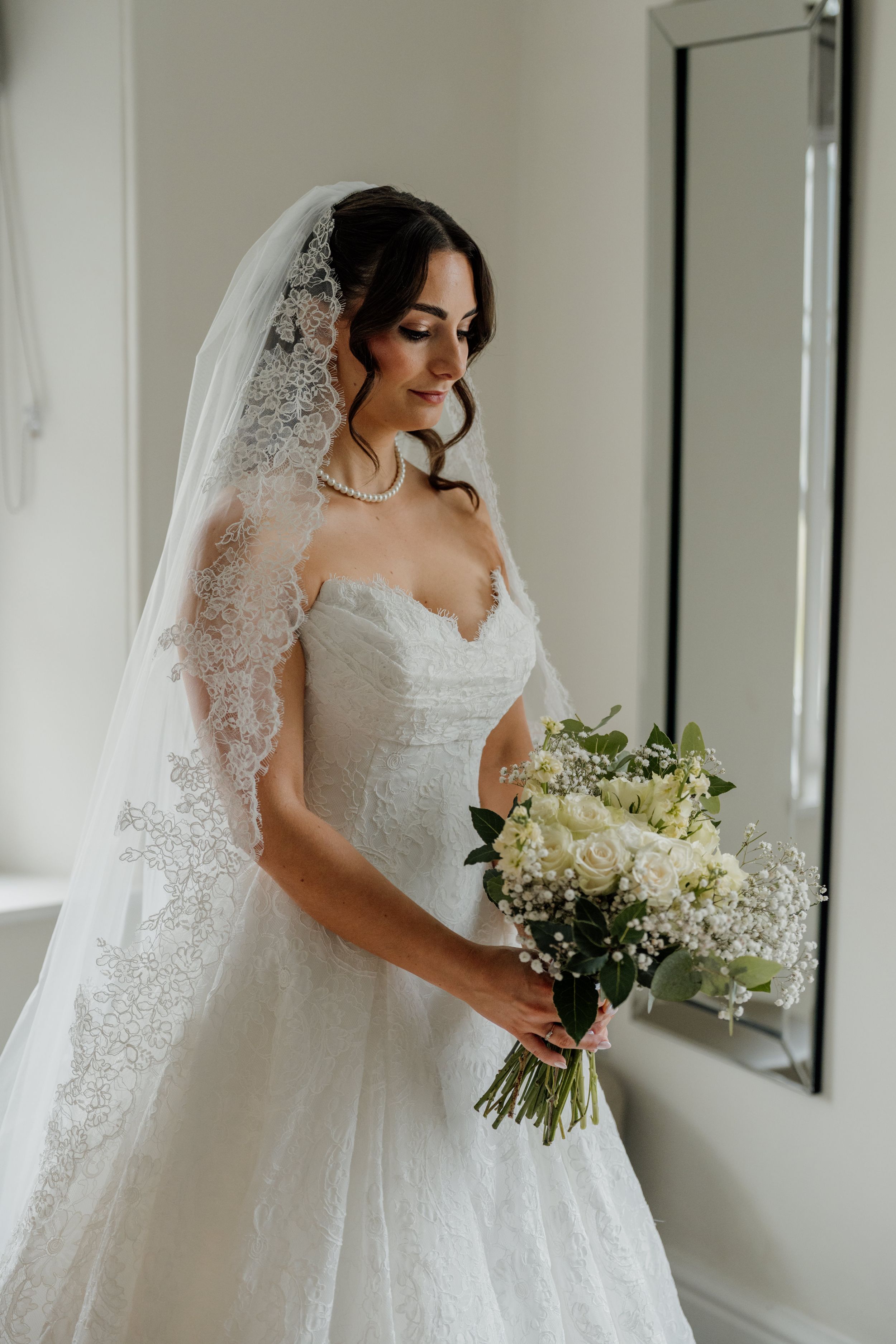 Bride in lace wedding gown and veil holding white rose bouquet indoors.