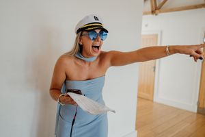 Woman in blue dress, captain hat, and sunglasses laughing and pointing at a party celebration.