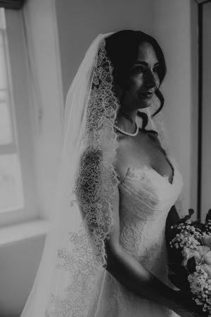 Elegant bride in lace wedding veil and strapless gown holding bouquet, black and white portrait photo.