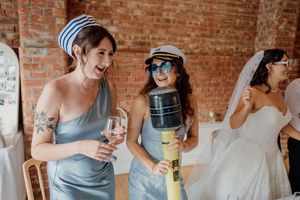Bridesmaids in blue dresses laugh and celebrate at a wedding reception, one holding wine, another with a toy hammer.