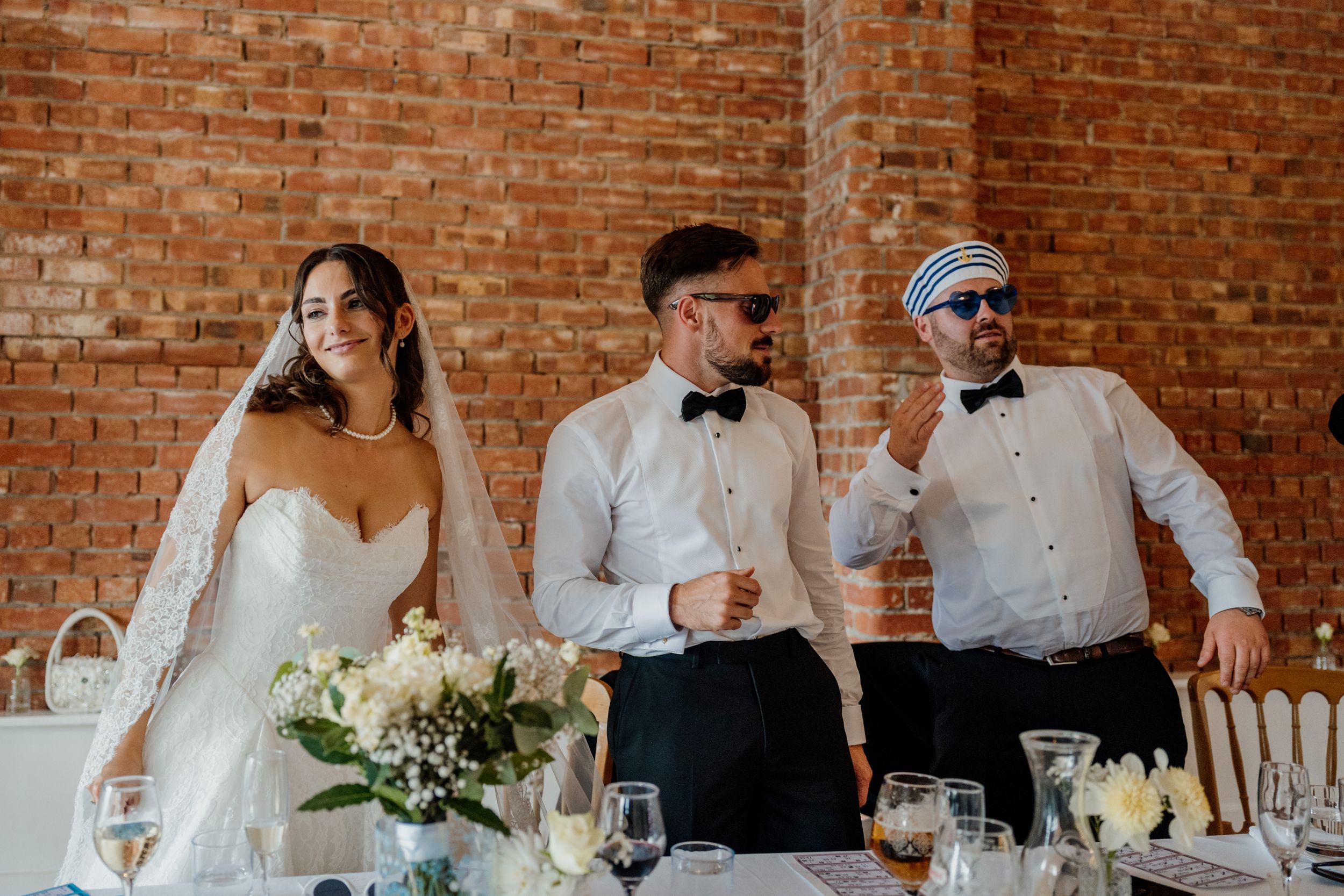Bride and two groomsmen in bow ties posing at wedding reception table with brick wall backdrop.