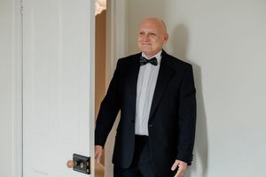 Elegant bald man in black tuxedo and bow tie standing in doorway at formal event.