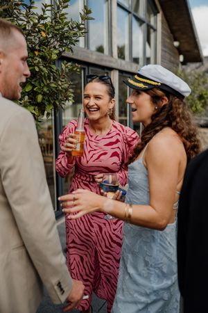 Wedding guests laugh and socialize outdoors, one wearing a pink zebra print dress and another a captain's hat.