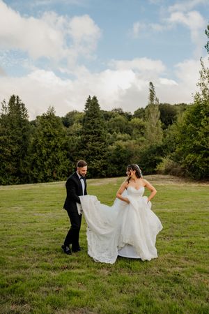 A groom in a black tuxedo lifts the train of his bride's white lace wedding dress on a green meadow.