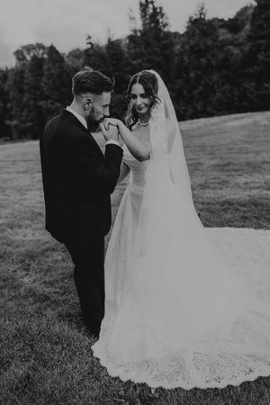 Black and white photo of a groom kissing his bride's hand on a lush green lawn.