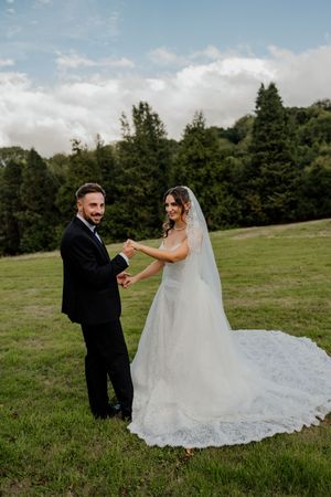 A smiling bride and groom hold hands in a lush green field on their wedding day.