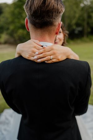 A bride with a diamond engagement ring embraces her groom from behind outdoors at their wedding.