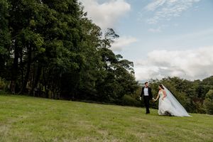 A bride and groom hold hands on a lush green hillside surrounded by tall trees under a partly cloudy sky.