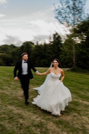 A joyful bride and groom run hand in hand across a green field in their wedding attire.