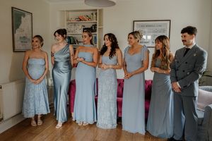 Bridesmaids in blue satin and lace gowns stand with a groomsman in a grey suit in an elegant room.