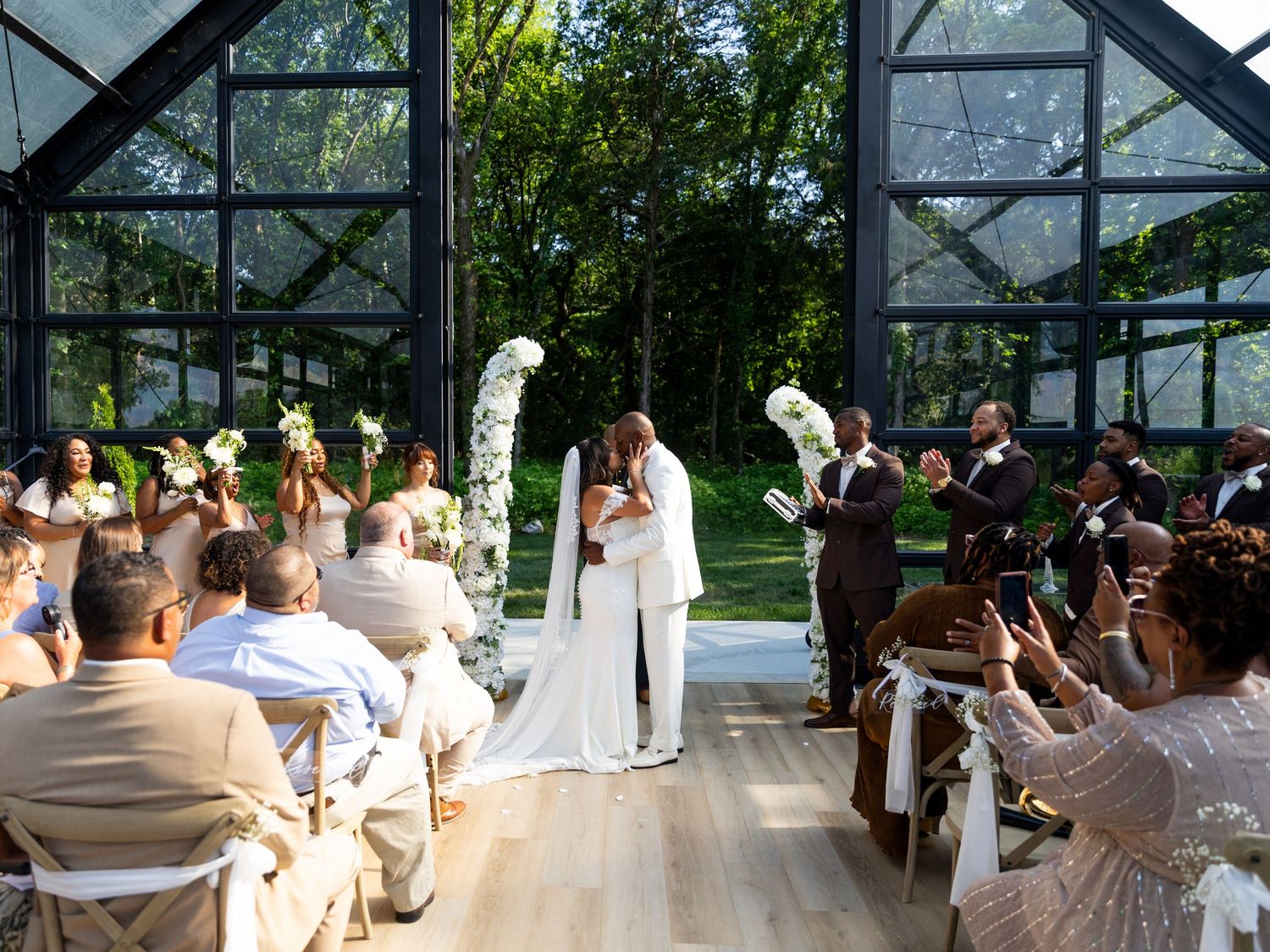 Couple shares first kiss at elegant greenhouse wedding ceremony with bridesmaids and guests celebrating in background.