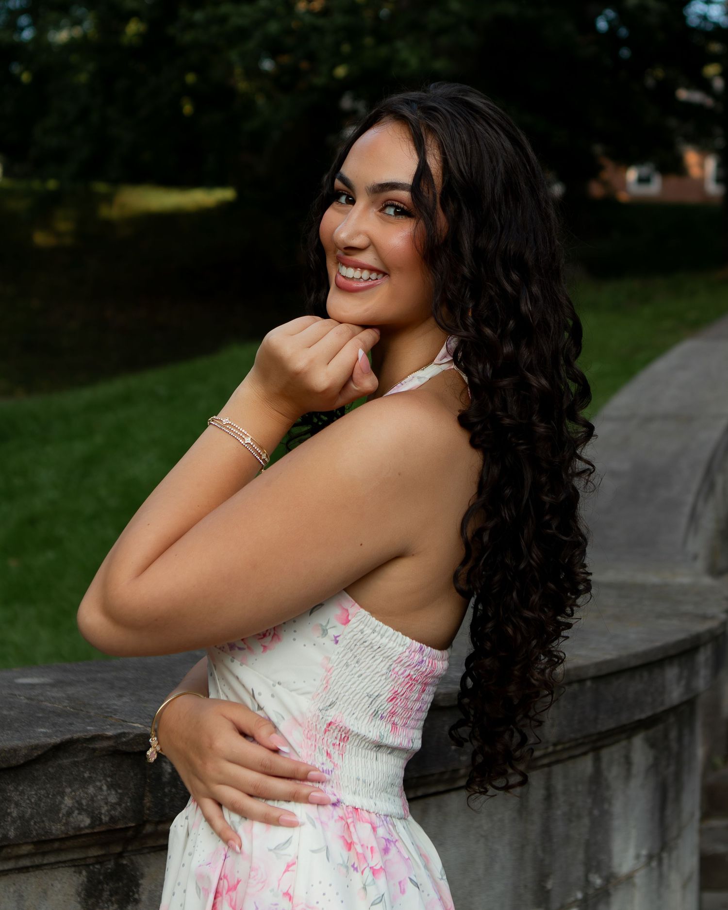 Young woman with long curly dark hair smiles over shoulder in floral dress, posing outdoors near stone railing.