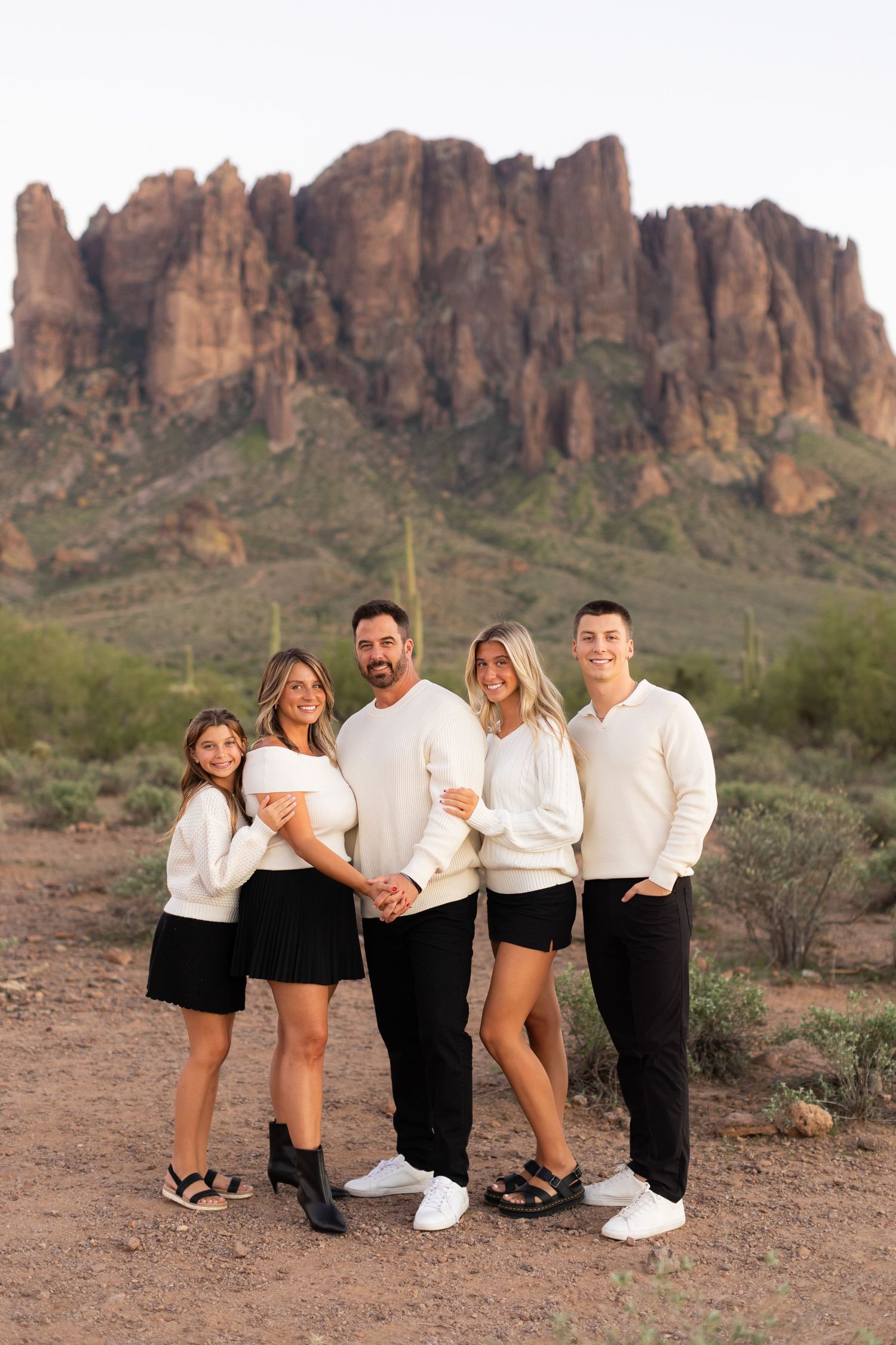 Family of five in matching cream tops and black bottoms posing before Superstition Mountains in Arizona desert.