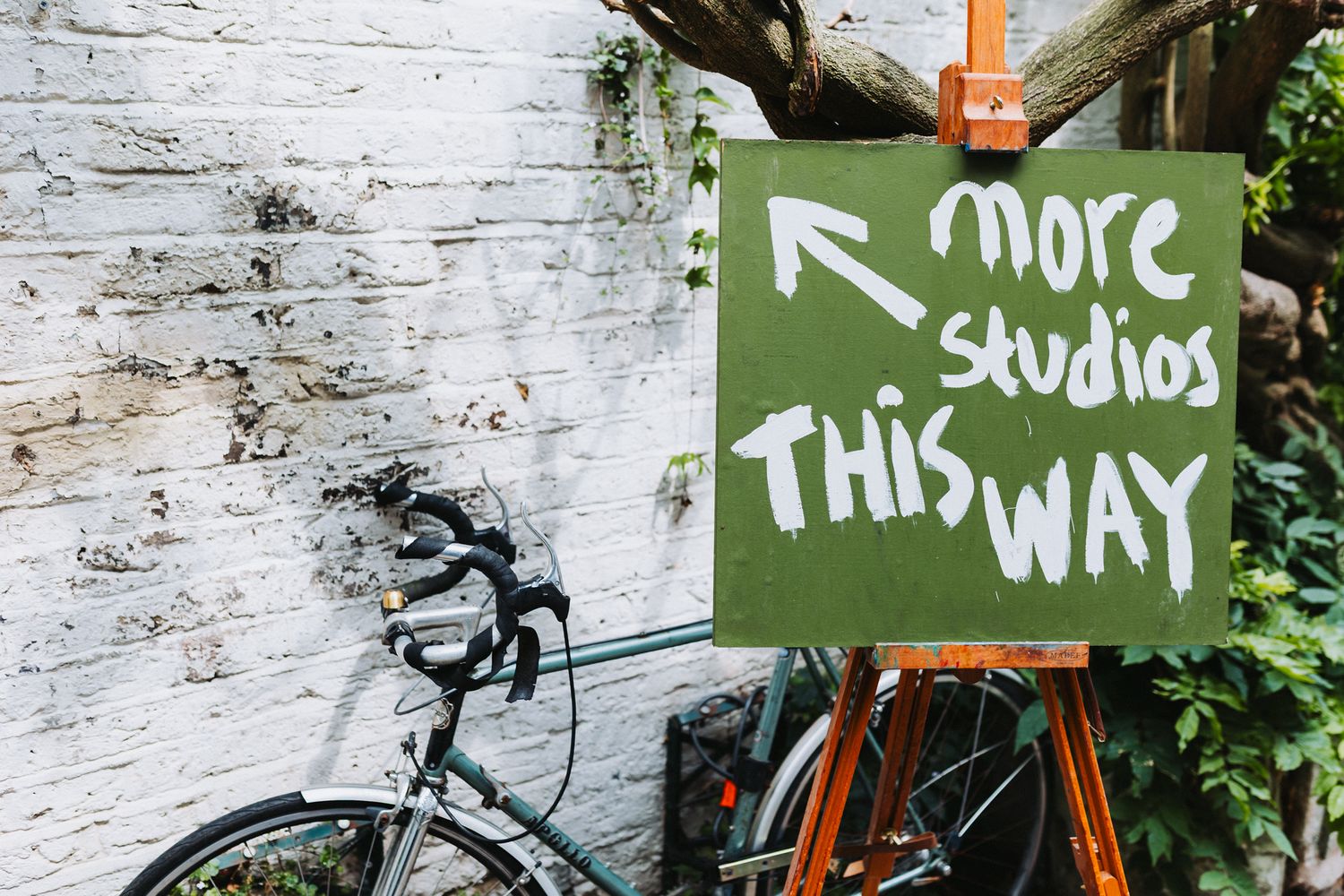 Green sign on easel reading 'More Studios This Way' with arrow, leaning against white brick wall beside a bicycle.