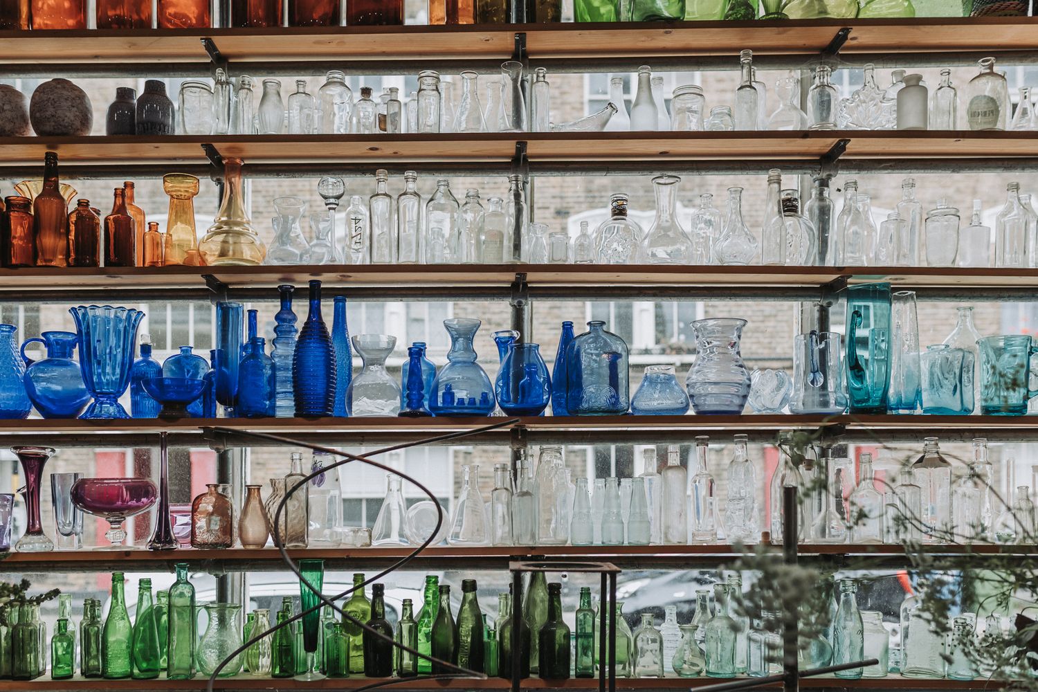 Colorful glass bottles and vases displayed on rustic metal shelves, featuring blue, amber, and clear glassware collections.