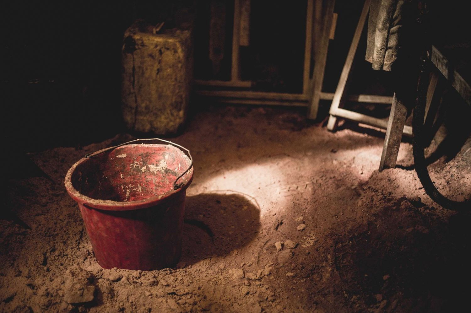 Old red bucket on a dirt floor in a dimly lit rustic wooden shed interior.