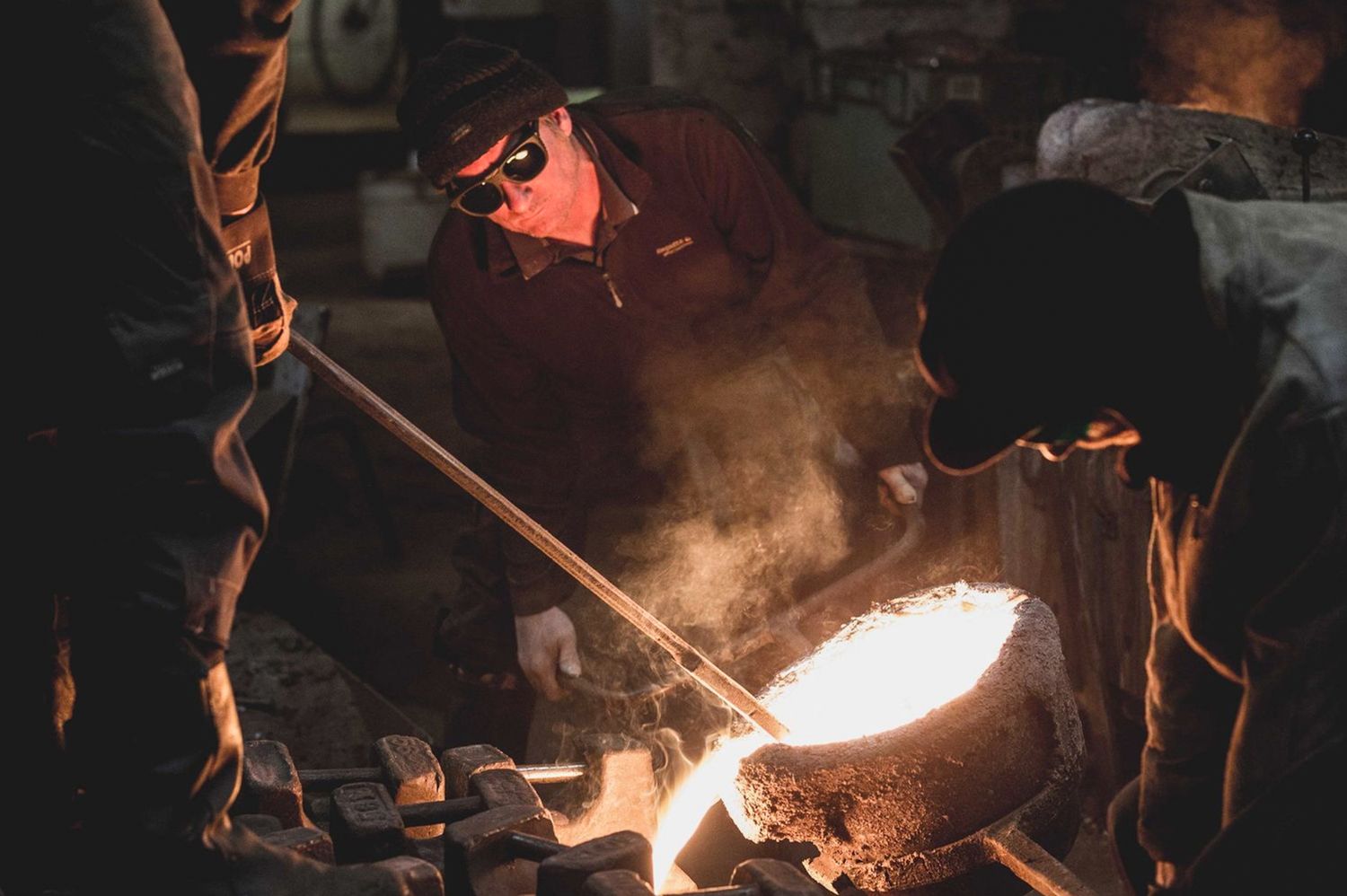 Workers in protective gear pour molten metal from a glowing crucible in a dark industrial foundry.